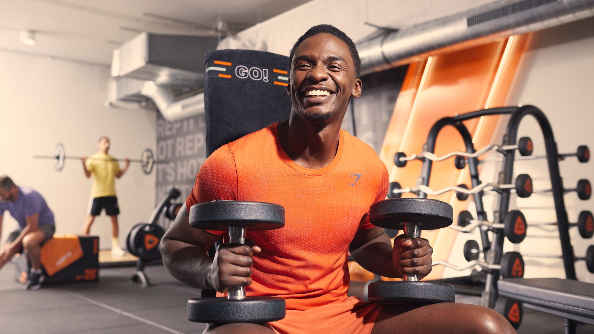 Un homme sourit, assis sur un banc dans une salle Basic-Fit, tenant deux haltères pendant un entraînement de musculation. D’autres adhérents s’entraînent en arrière-plan parmi les équipements de musculation et des touches orange.